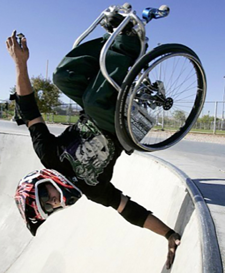 A wheelchair rider goes fully inverted at a skate park — chair overhead, arms extended, helmet on
