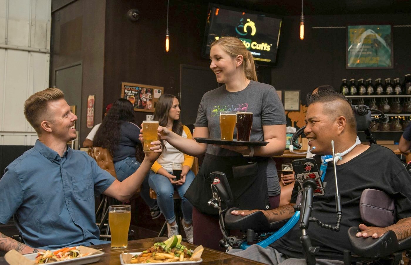 A server carrying a tray of beers smiles as she hands a glass to a man at the table, while another man in a power wheelchair with breathing equipment smiles along, at a busy pub.