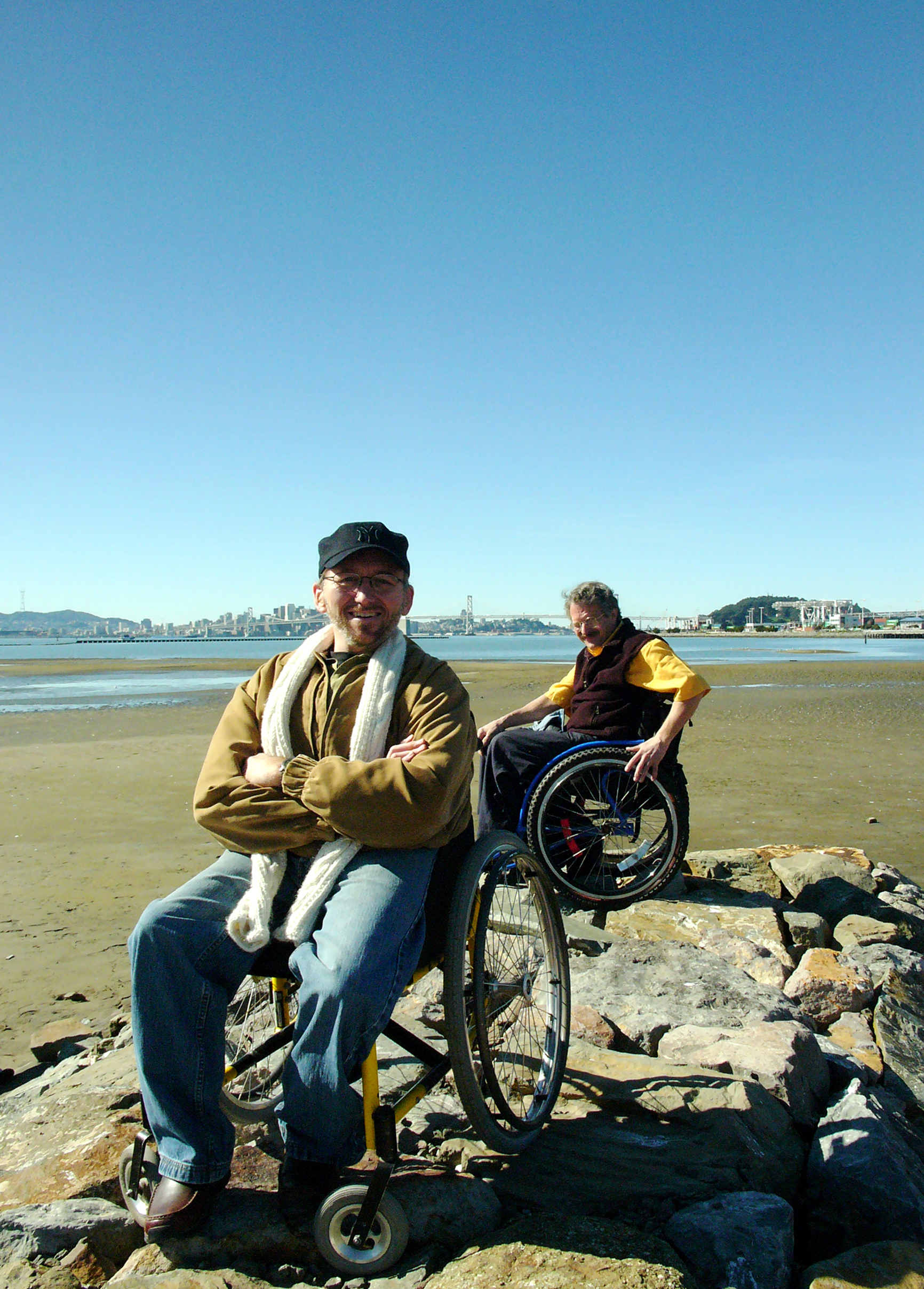 Erick and a friend, both in wheelchairs, on rocks at Alameda Beach with the Bay Bridge behind them