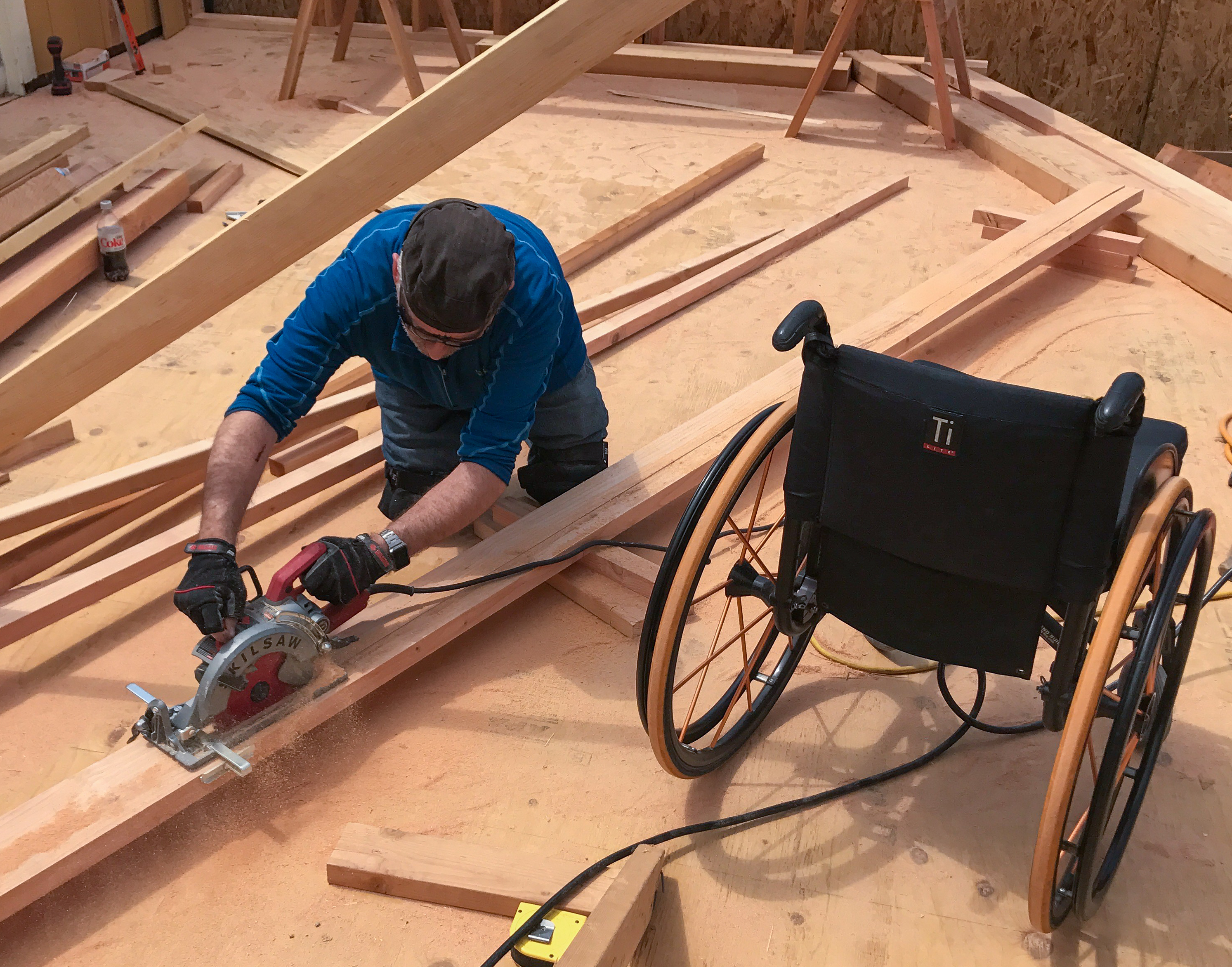 Erick using a circular saw on a construction site, his wheelchair right beside him